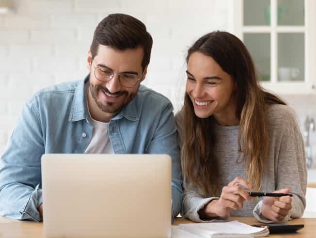 Young couple sitting at dining table looking at laptop
			