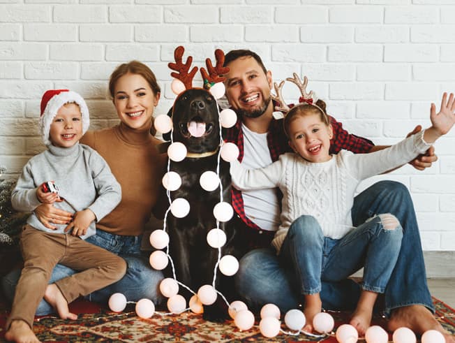 Hispanic couple with two young children. Christmas lights are draped around them and their dog,